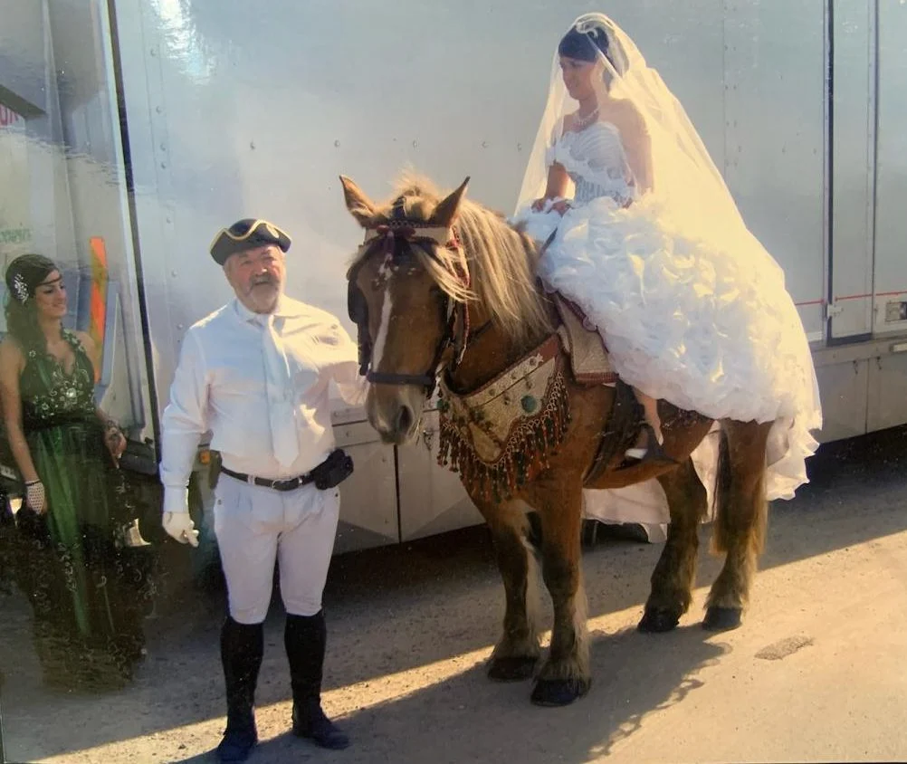 Mariée montée en amazone sur un cheval de trait, selle arabe brodée, accompagnée du cocher en tenue