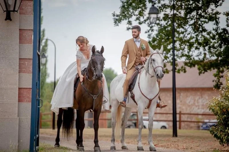Les mariés à cheval côte à côte devant les écuries, la mariée en robe longue